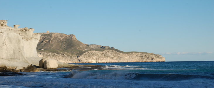 Wellen rollen an den Strand, im Hintergrund ein etwa 50 Meter hoher Felsen unter wolkenlosem Himmel