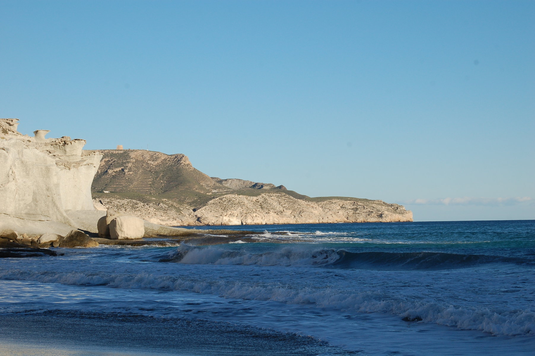 Wellen rollen an den Strand, im Hintergrund ein etwa 50 Meter hoher Felsen unter wolkenlosem Himmel