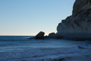 Ein Felsen im Meer an den die Brandung schlägt unter blauem Himmel.