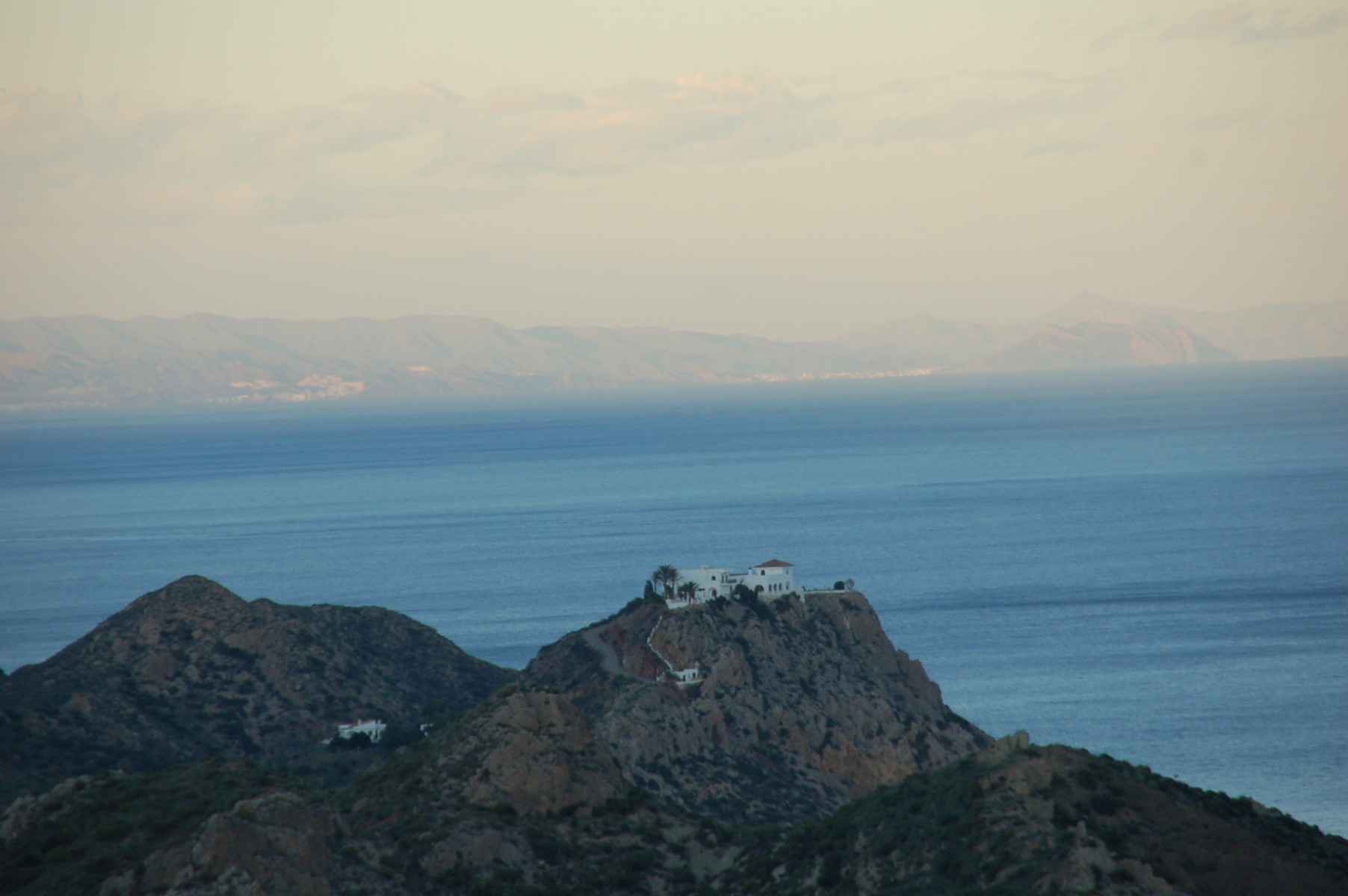 Man sieht im Abendhimmel ein einsames typisch für Andalusien weiß getünchtes Haus in einer schroffen felsigen Umgebung, im Hintergrund das Mittelmeer