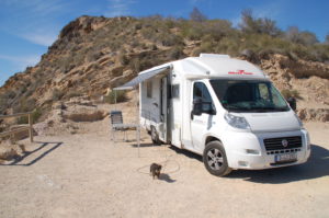 Das Wohnmobil steht einsam und allein auf dem Parkplatz der Batteria de Castillitos. Das Sonnensegel ist ausgefahren, ein Stuhl und ein Tisch stehen darunter. Die Landschaft ist mediterran karg. Der Dackel steht neben dem Wohnmobil und blickt in die Kamera