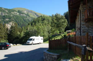 Das Wohnmobil ist am Waldrand geparkt. Der Untergrund ist schräg, weshalb die Vorderräder auf Plastikrampen stehen, um das Gefälle auszugleichen. Im Hintergrund sieht man die Schweizer Alpen.